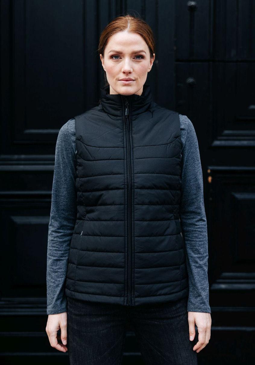 Woman in a black quilted vest over a grey top against a dark door.