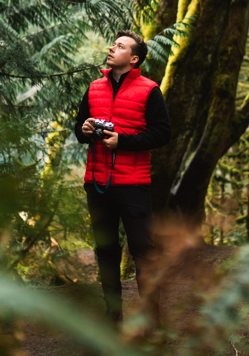 Man in a red quilted vest holding a camera in a green forest.