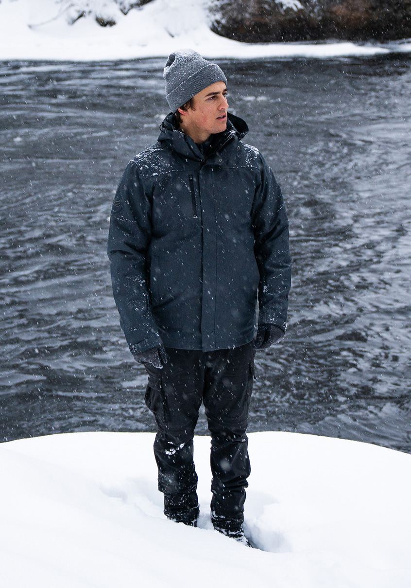 Man in a grey jacket standing in snow by a river.